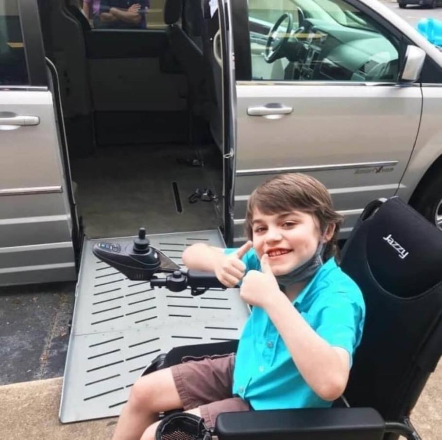 A child gives a thumbs-up sign from his wheelchair alongside a side entry wheelchair van with a ramp.